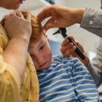 Avaliação de otite média infantil: profissional de saúde examina a orelha de uma criança com otoscópio, com responsável ao lado durante a consulta.