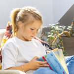 Young girl with microtia wearing a cochlear implant sits on a chair reading a book, showing how children with hearing loss can enjoy learning and everyday activities.