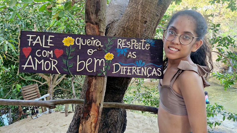 Clara com implante coclear ao lado de uma placa com as frases Fale com Amor, Queira o Bem e Respeite as Diferenças