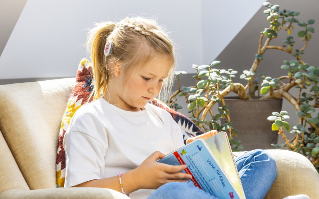 Menina com microtia usando um implante coclear sentada em uma cadeira e lendo um livro, mostrando como crianças com perda auditiva podem aproveitar a aprendizagem e as atividades do dia a dia.