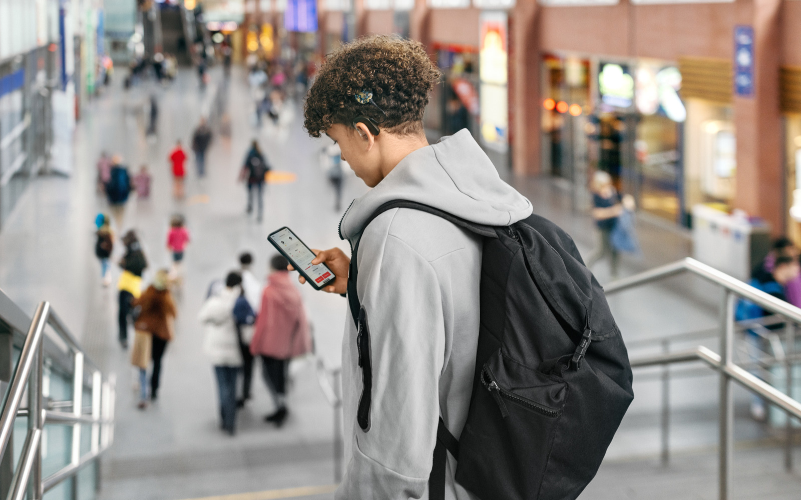 A teenager with a cochlear implant looks at his phone. The setting is a railway station.