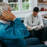 Homem mais velho sentado leva a mão ao ouvido enquanto conversa com um profissional de saúde durante uma avaliação auditiva.