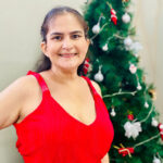 Karine usando vestido vermelho, com árvore de Natal ao fundo.