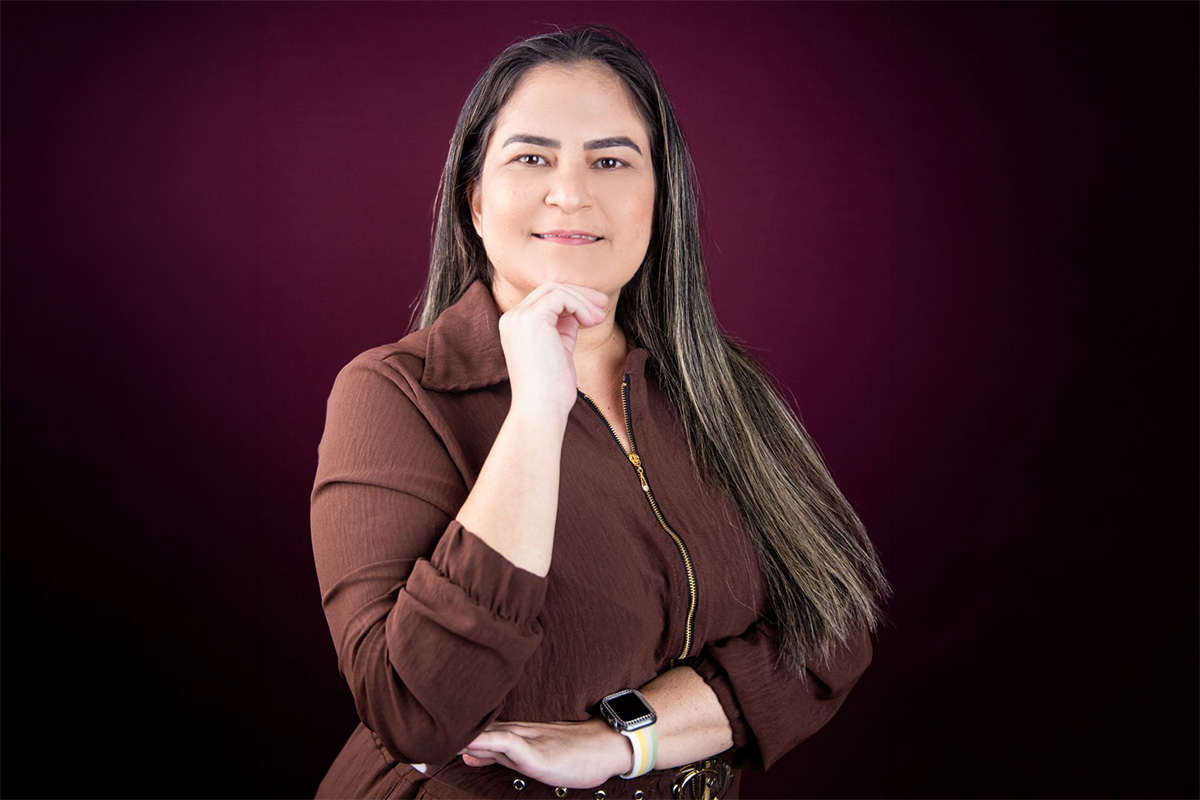 Karine em fundo liso escuro em pose profissional com a mão apoiada no queixo.