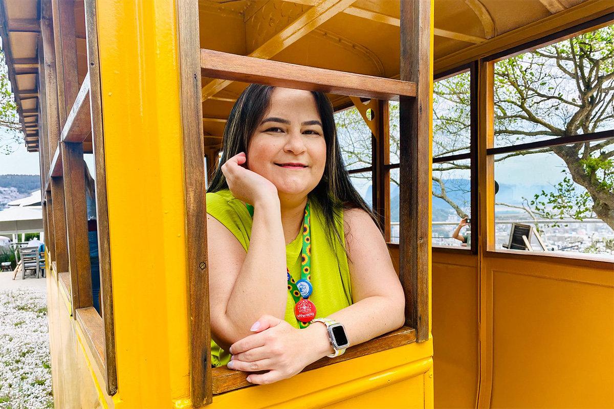 Karine apoiada na janela de um vagão amarelo em área externa, com paisagem ao fundo.