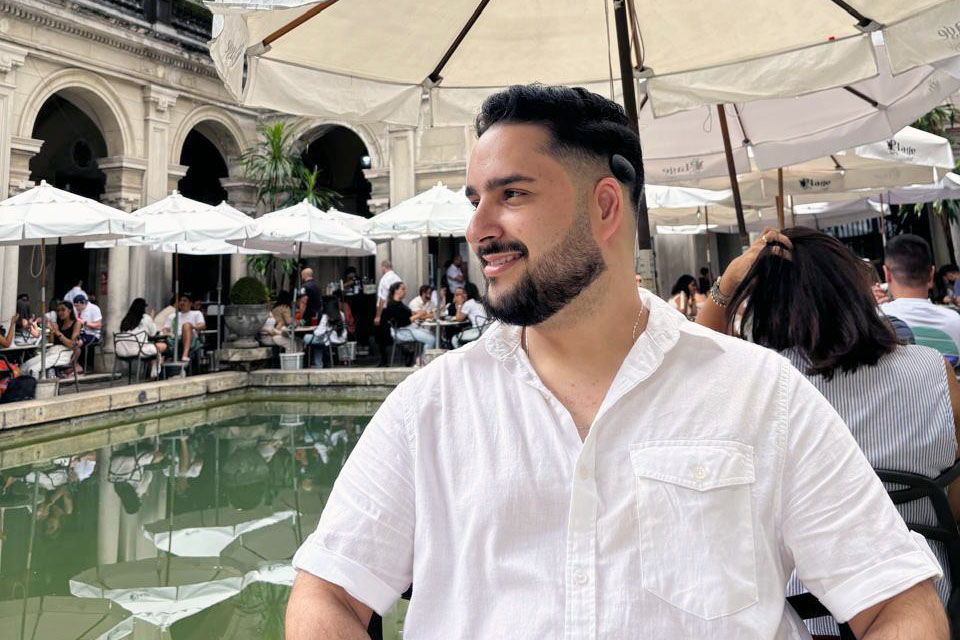 Arthur sentado em área externa de café, usando camisa branca, com espelho d’água à frente e mesas com guarda-sóis ao fundo.
