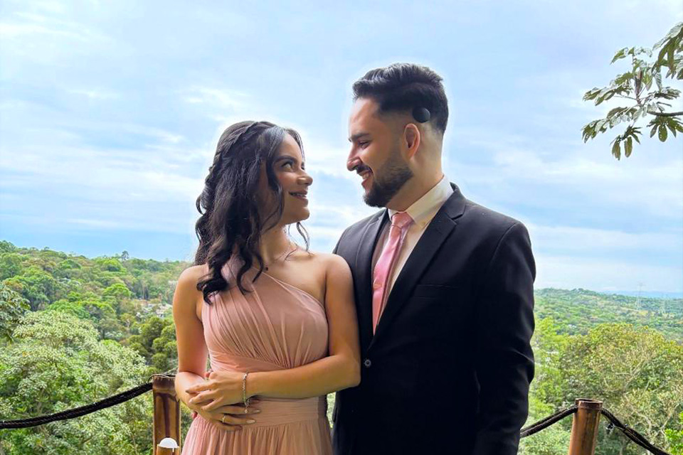 Arthur e sua esposa em trajes formais, de pé em varanda com vista para área verde; ela usa vestido rosa e ele, terno escuro com gravata rosa.
