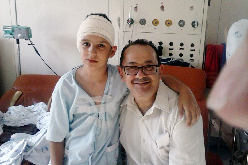 Arthur sentado em ambiente hospitalar com avental médico e curativo na cabeça.