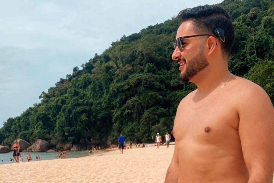 Arthur em pé na areia da praia e de óculos escuros.