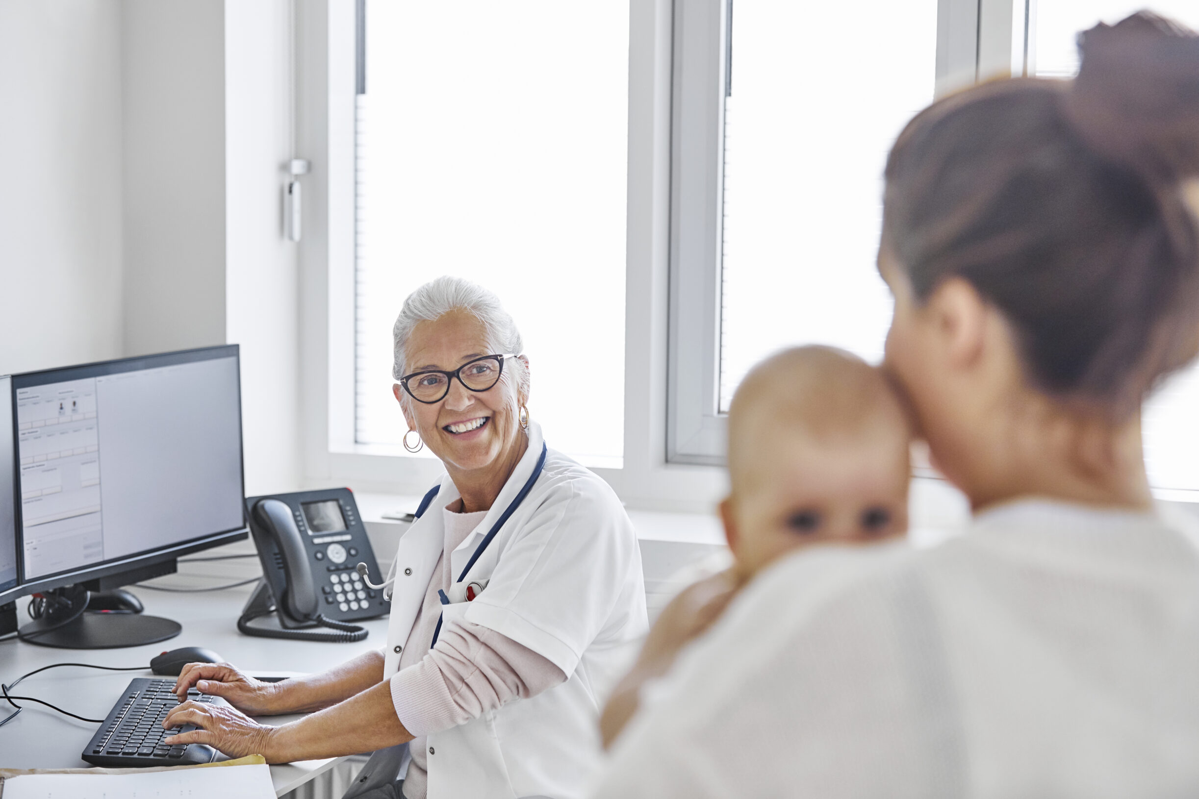 Profissional de saúde em consultório conversa com responsável que segura um bebê durante atendimento pediátrico.