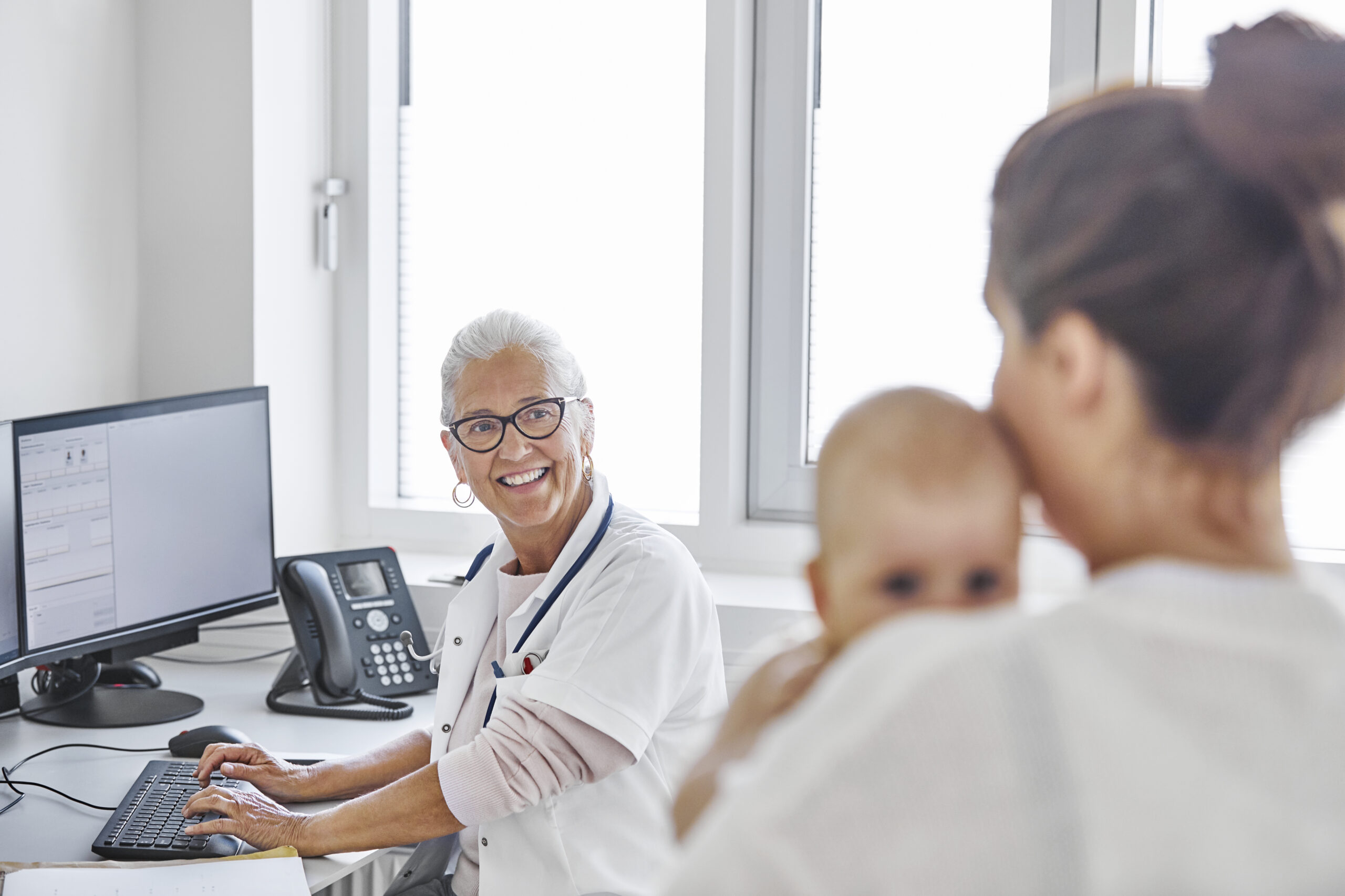 Profissional de saúde em consultório conversa com responsável que segura um bebê durante atendimento pediátrico.