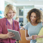 Profissional de saúde mostra informações em tablet a responsável com bebê em ambiente clínico.