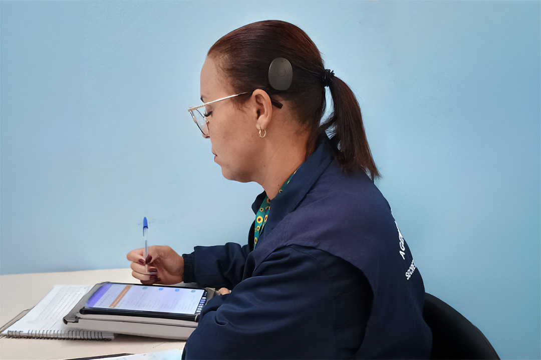 Maria Angélica utiliza implante coclear enquanto faz anotações e consulta um tablet durante atividade profissional.